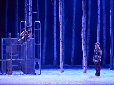 An actress perches atop a metal frame. She gazes down softly at an actor wearing a hat. The lighting is blue and behind them are tall birch trees.