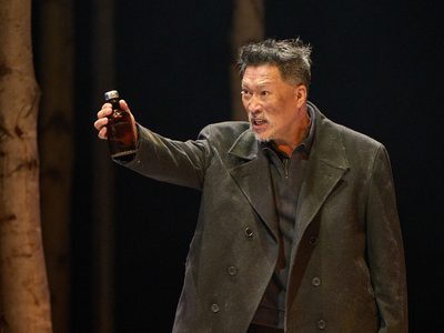 A middle aged actor wears a well-worn black coat. He has an angry facial expression. He holds up a vial of an unknown substance.