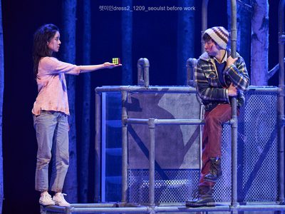 An actor and and actress stand on metal frames.  The actress wears a pink top and casual jeans. She holds a Rubik's cube out to the actor who seems both impressed and surprised.