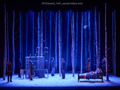 Seven actors are dotted about the stage, they look up at the sky as snow falls down. two other actors are in bed, one sleeps while the other gazes down at her.