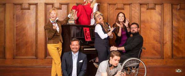 A group of people are on a stage. They are surrounding a piano with an award on top. At the front, a man on the left wearing a suit sits up straight with his hands clasped and smiles at the camera. Behind him, a woman in yellow trousers and brown suit jacket has one hand on the award on top of the piano and is looking towards to the camera, smiling. Sitting on top of the piano is a young woman in a red and white tracksuit with black stripes. She has her feet on the piano and one arm raised in a pose in the air. In front of the piano is a woman in formal office wear, throwing her head back and laughing. Next to her is a woman is a fuschia top doing jazz hands, and a man with a beard and glasses wearing a turtle neck is sitting in a wheel chair. They are all smiling and looking directly at the camera. At the foot of the stage on the left is a young man wearing a grey top and dark trousers - he is texting on his phone.