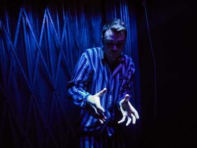 A man stands in front of a dark blue background with his hands gesturing in front of him.
