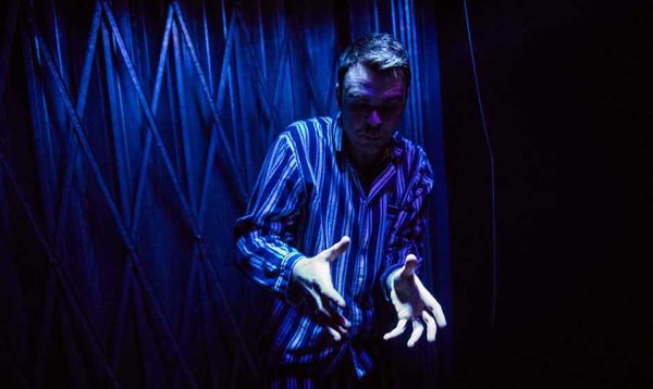 A man stands in front of a dark blue background with his hands gesturing in front of him.