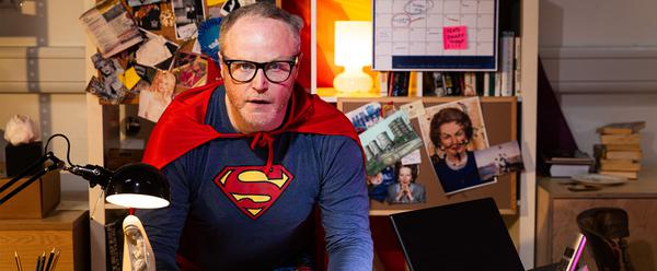 DB Dressed as superman leaning over a desk. behind him is a bulletin board which prominently features Margaret thatcher