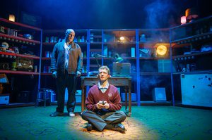 Production imageof a scene from Maggie & Me. DB is standing behind Mark, who sits in the spotlight on the ground, legs in a basket.