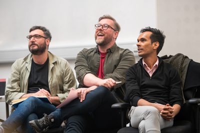 Three men are sitting in chairs watching rehearsals, one of them is laughing.