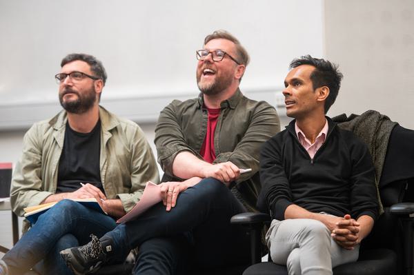 Three men are sitting in chairs watching rehearsals, one of them is laughing.