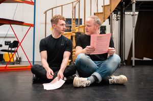 Two men sit on the ground with their legs crossed, looking at one another, holding their scripts.
