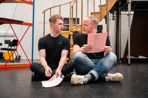 Two men sit on the ground with their legs crossed, looking at one another, holding their scripts.