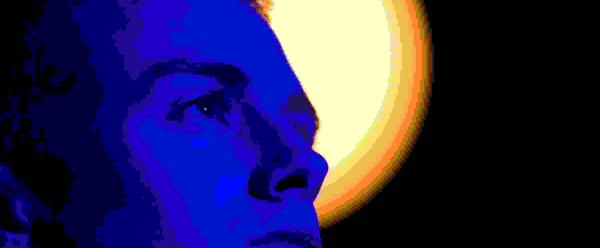 A young man stands looking upwards. He is bathed in a blue light. Behind him against a black backdrop, an circular yellow light is illuminated.