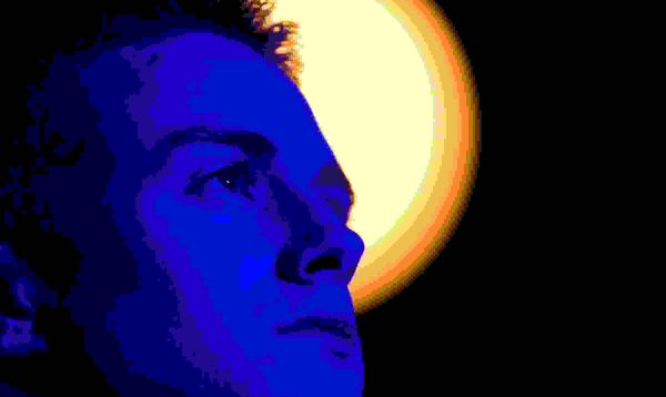 A young man stands looking upwards. He is bathed in a blue light. Behind him against a black backdrop, an circular yellow light is illuminated.