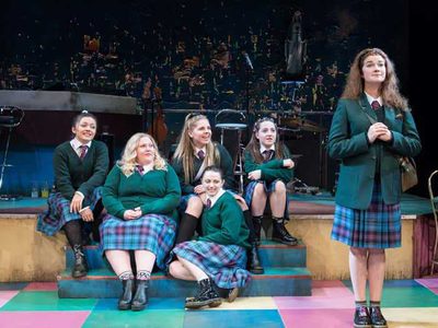 A group of five girls in school uniform giggle as they watch as a classmate stands obediately at the front of the stage