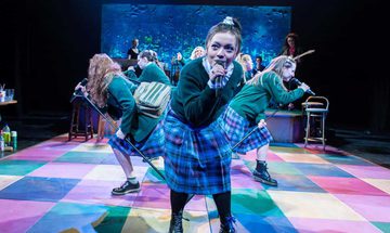 A group of schoolgirls in uniform lean into microphones as they sing on a colourfully tiled stage