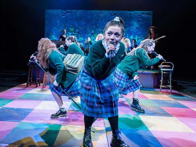A group of schoolgirls in uniform lean into microphones as they sing on a colourfully tiled stage