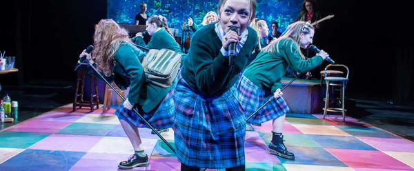 A group of schoolgirls in uniform lean into microphones as they sing on a colourfully tiled stage