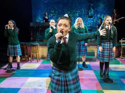 School girls dressed in green jumpers and tartan skirts sing into microphones, a band playing in the background.