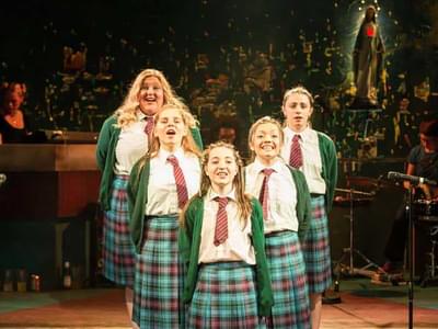 Girls in school uniform stand in a v shape, singing with their arms behind their backs