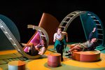 Alt text: Three performers on a brightly coloured stage interact with large curved set structures resembling tunnels or climbing frames; one crouches at centre while two recline on circular platforms, dressed in school-style costumes under theatrical lighting.