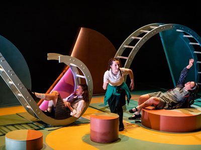 Alt text: Three performers on a brightly coloured stage interact with large curved set structures resembling tunnels or climbing frames; one crouches at centre while two recline on circular platforms, dressed in school-style costumes under theatrical lighting.