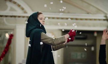 A woman in a travelling cloak mines driving a car as fake snow flies around her
