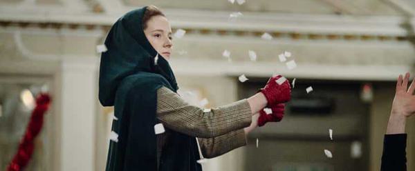 A woman in a travelling cloak mines driving a car as fake snow flies around her