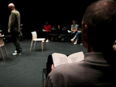 An audience member is visible from the book, from the back. There are several other audience members in the back. Tim Crouch stands on stage with two chairs on the left hand side of the image.