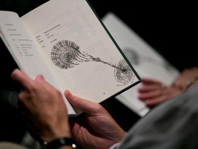 An audience member holds the book open, the page has text and drawing by Rachana Jadhav.