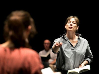 Shyvonne Ahmmad and Susan Vidler face each other, both holding a book. Shyvonne Ahmmad is seen from the back, in the foreground, and Susan Vidler is pointing in her direction. Some audience members are visible in the foreground.