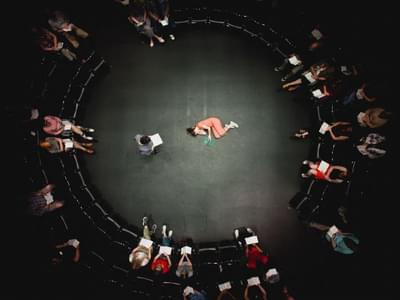 The photograph is taken from above, and looks down on a circle made off of two rows of chairs, with the stage in the enclosed circle. Shyvonne Ahmmad is seen lying down on the floor wearing orange, holding a book with a green cover. Susan Vidler is seen standing up holding a book on the left. Audience members are sat on chairs.