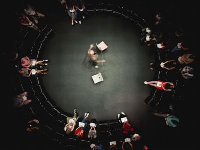 The photograph is taken from above, and looks down on a circle made off of two rows of chairs, with the stage in the enclosed circle. Tim Crouch is seen standing up in the centre of the stage, two chairs each side of him. Audience members are sat on chairs.