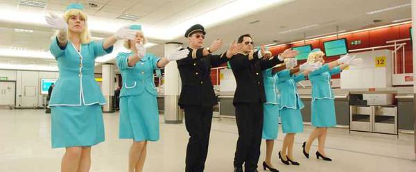 In an airport check in lounge, five female air stewards dressed in turquoise and two male pilots dress in black wearing sunglasses stand in a line. All have their arms raised in front of them, their hands flat and gesturing forwards.
