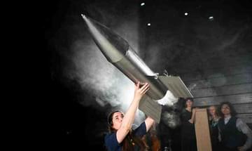 A woman holds a large silver rocket above her head as it prepares to fly