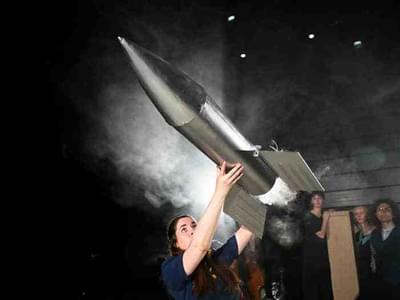 A woman holds a large silver rocket above her head as it prepares to fly