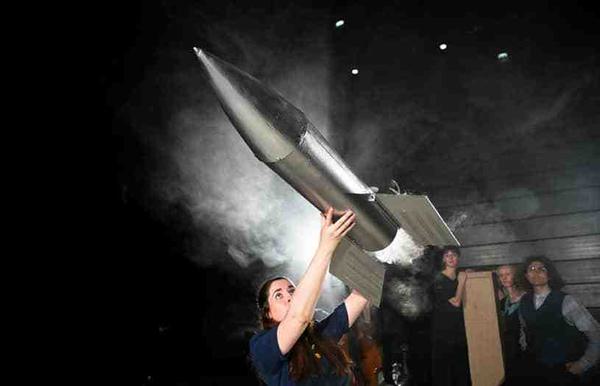 A woman holds a large silver rocket above her head as it prepares to fly