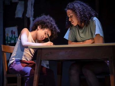 A little boy writes at a desk as his mum looks on.