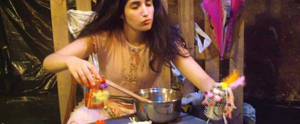 A young woman with long dark hair sits on the ground. In front of her on a table is a saucepan and a can of fizzy juice. She holds two cans of fizzy juice in her hands.