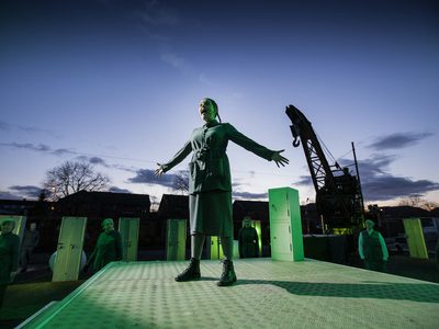 A woman in overalls stands with her arms wide. The shadow of a crane behind her.