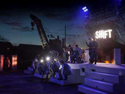 A man in a flat cap holds a steel mug that says "Shift". A crane is behind him.