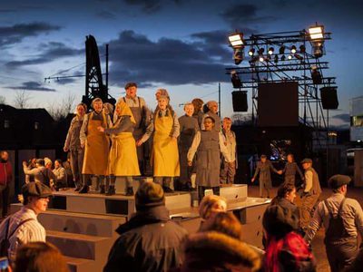 A group of people in overalls stand in a crowd with an industrial crane in the background