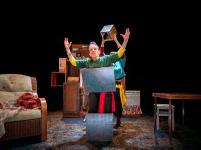A woman is standing inside a silver box, with another box around her chest and her arms raised above her, with a snarl on her face. Behind her, someone is holding a small box above her head.