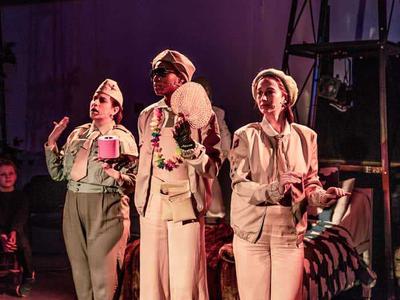 The three slumber sisters standing in a row. Augusta in the centre is wearing sun glasses and has a wreath round her neck. She is holding a fan and a pink toilet roll.