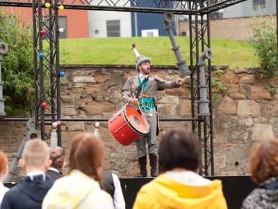 A man with a periscope on his head and a drum stands on a stage outside