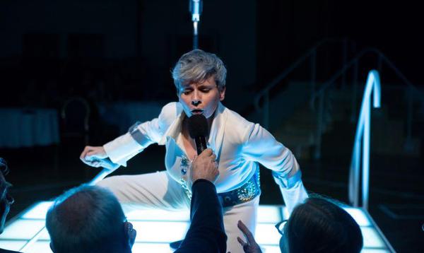 Claire Cunningham is seen in an Elvis Presley white costume, singing in a microphone handed by a member of the audience, while crouching on a table.