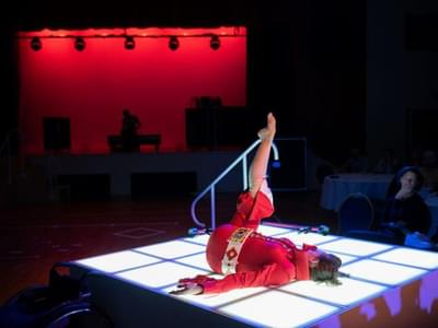 Tanja Erhart is seen on a lighted stage in a red Elvis Presley costume, lying on the floor one leg up.