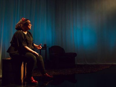 Hannah Lavery is sitting on a box. She is all dressed in black with red shoes and a red hairband visible. The background in dark blue. She is seen talking, facing to the right of the frame.