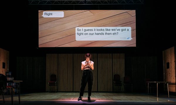 A woman stands looking at her phone, text messages are projected on a screen behind her