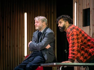 Two men, one sat on a table, one leaning over it stare forwards