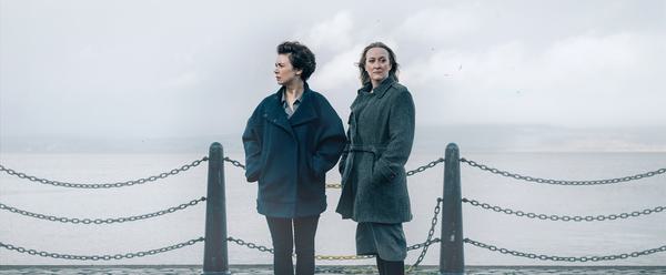 Two women stand, windswept against a cloudy backdrop
