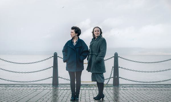 Two women stand, windswept against a cloudy backdrop