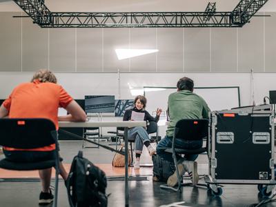 An actor sits at the front of the space delivering a script whilst other production members look on